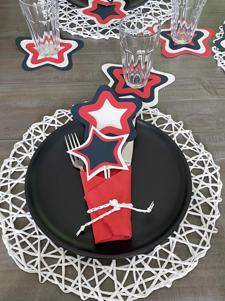 A table setting with a black plate on a white woven placemat, topped with a red napkin, silverware, and layered red, white, and blue star-shaped decorations—perfect for Patriotic Entertaining Ideas. Clear glasses and more star decor complete the festive table.