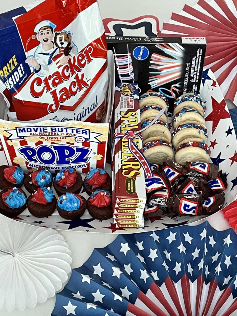 A patriotic-themed snack tray, perfect for Patriotic Entertaining Ideas, features Cracker Jack, Popz popcorn, Sparklers, cupcakes with red, white, and blue icing, cookies, and chocolate candies surrounded by American flag decorations.