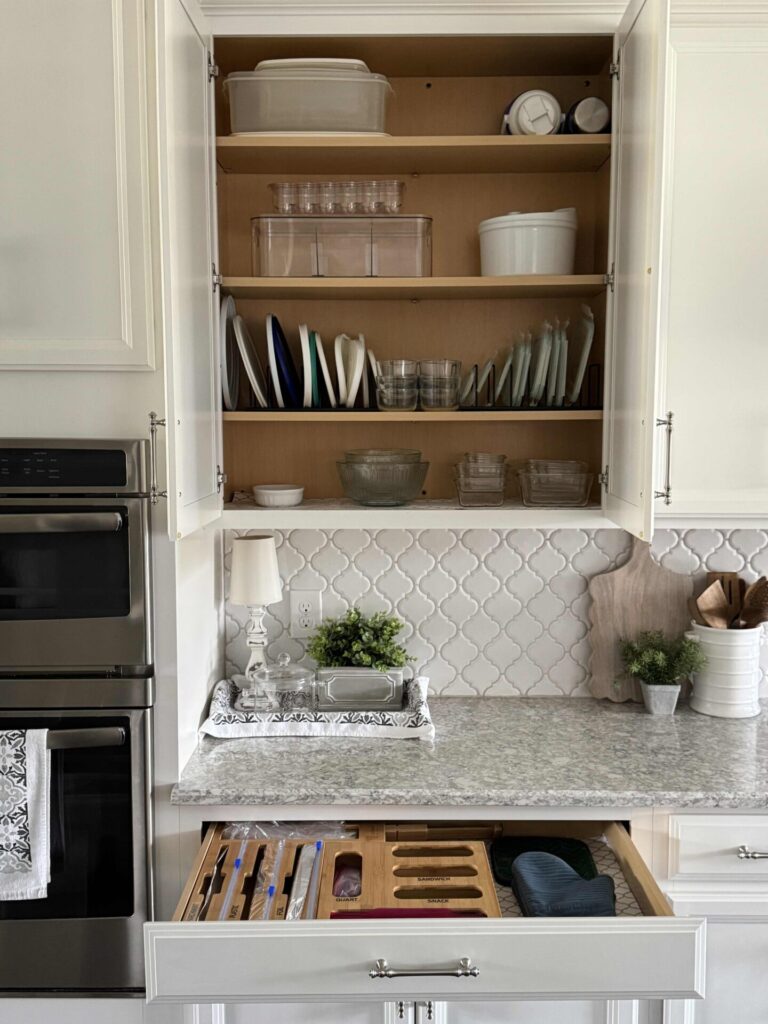 A neatly organized kitchen with open cabinets displaying plates, bowls, and containers, and an open drawer with utensils. The counter holds a tray with a lamp, plants, cutting boards, and space to organize greeting cards against a tiled backsplash.