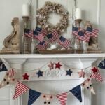 A white fireplace mantel decorated with small American flags in glass bottles, rustic wood accents, a neutral wreath, and a red, white, and blue star and pennant garland offers festive Patriotic Entertaining Ideas.