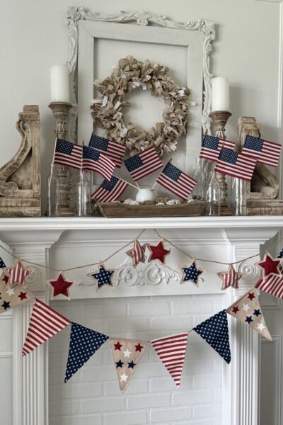 A white fireplace mantel decorated with small American flags in glass bottles, rustic wood accents, a neutral wreath, and a red, white, and blue star and pennant garland offers festive Patriotic Entertaining Ideas.