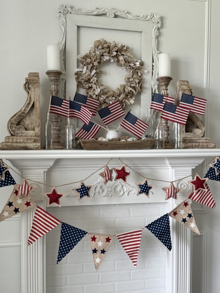 A white fireplace mantel decorated with small American flags in glass bottles, rustic wood accents, a neutral wreath, and a red, white, and blue star and pennant garland offers festive Patriotic Entertaining Ideas.