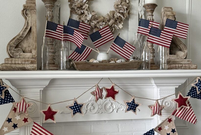 A white fireplace mantel decorated with small American flags in glass bottles, rustic wood accents, a neutral wreath, and a red, white, and blue star and pennant garland offers festive Patriotic Entertaining Ideas.