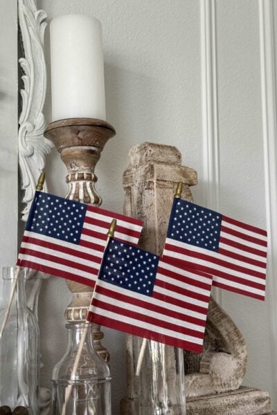 Three small American flags are displayed in clear glass bottles on a shelf, next to vintage-style wooden candle holders and a white candle—a charming touch for patriotic entertaining ideas against a light-colored wall with decorative molding.