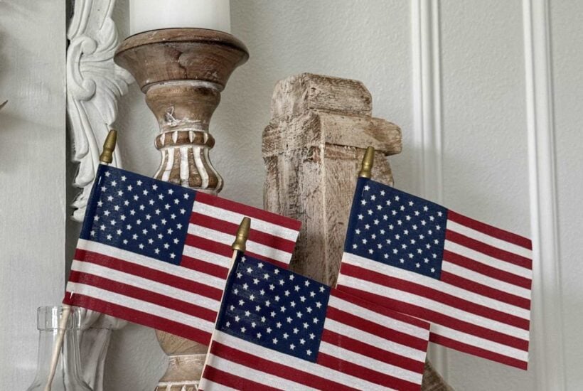 Three small American flags are displayed in clear glass bottles on a shelf, next to vintage-style wooden candle holders and a white candle—a charming touch for patriotic entertaining ideas against a light-colored wall with decorative molding.