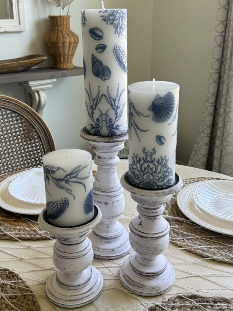 Three white pillar candles with blue seashell and coral designs are displayed on distressed white candle holders, perfect for patriotic entertaining ideas on a dining table with woven placemats, white plates, and a wicker vase in the background.
