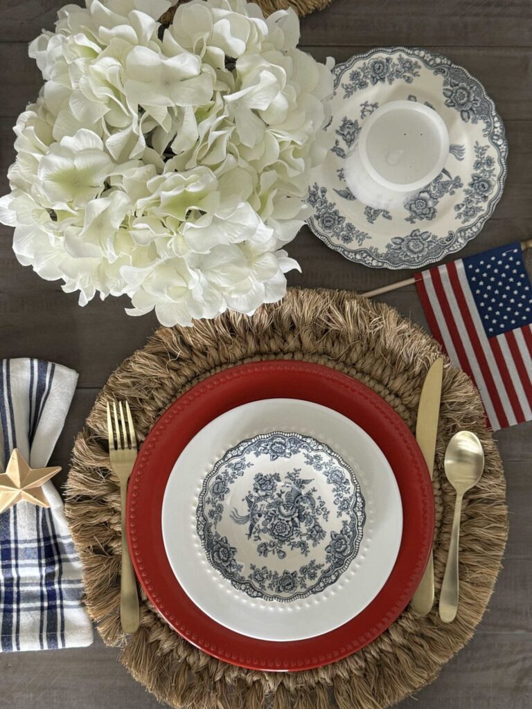 Overhead view of a table setting with gold utensils, a red plate, blue and white floral dishes, a woven placemat, patriotic entertaining ideas like a blue plaid napkin with a wooden star, white hydrangeas, and an American flag on a gray table.
