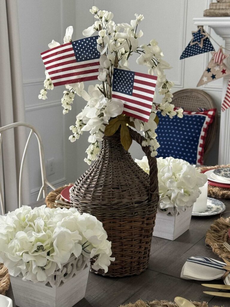 A woven basket vase with white flowers and two small American flags sits on a table, flanked by white flower arrangements—perfect for Patriotic Entertaining Ideas. Festive decorations and a blue pillow with white stars complete the scene.