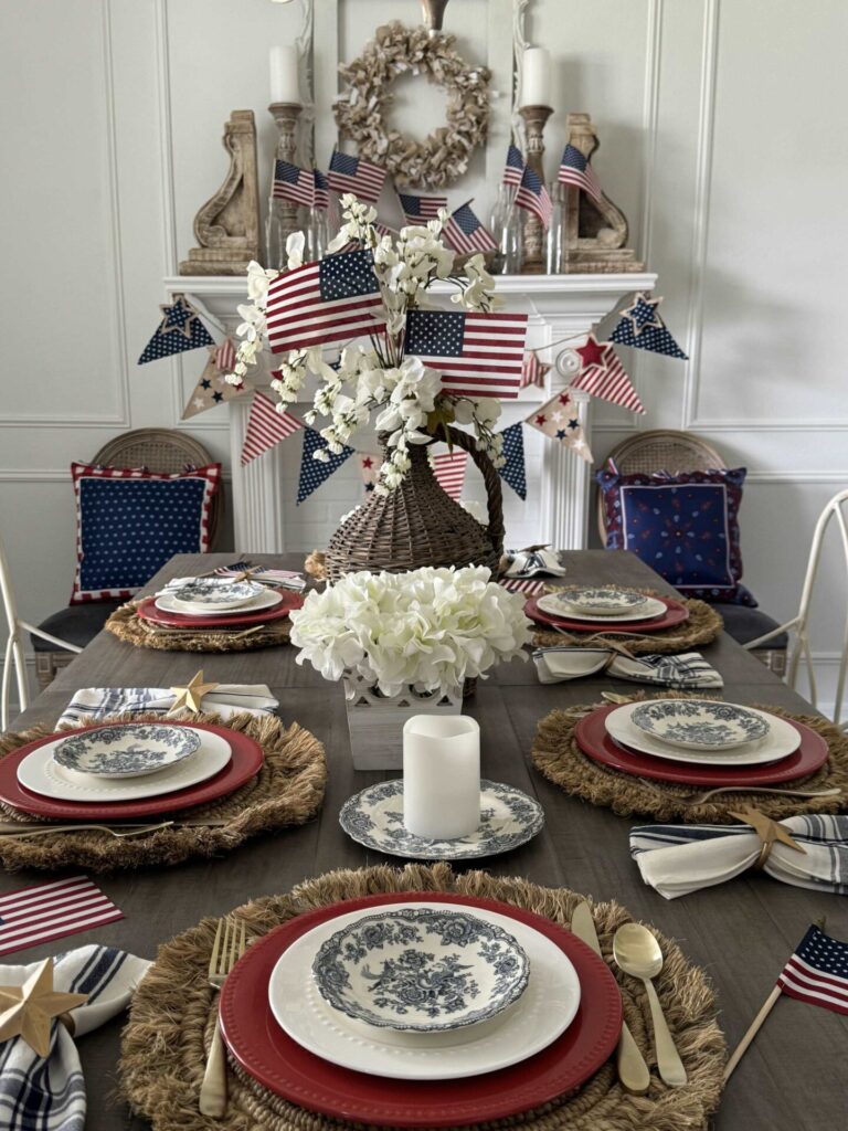A dining table set for a patriotic celebration showcases Patriotic Entertaining Ideas with American flag decorations, blue and white floral-patterned plates, red chargers, woven placemats, white flowers, candles, and a mantel adorned with flags and pillows.
