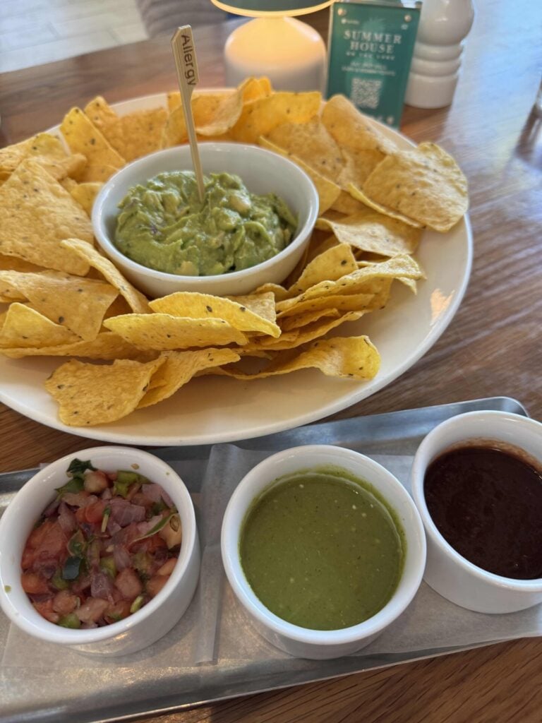 A plate of tortilla chips with guacamole in the center is served alongside three small bowls of salsa: chunky pico de gallo, green salsa, and dark red salsa, all on a wooden table.