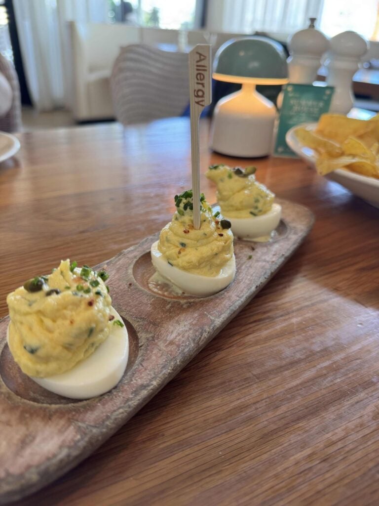 Three deviled eggs topped with herbs are arranged on a wooden serving tray. One egg has a stick labeled Allergy. In the background, there are chips, salt and pepper shakers, and a softly lit lamp.