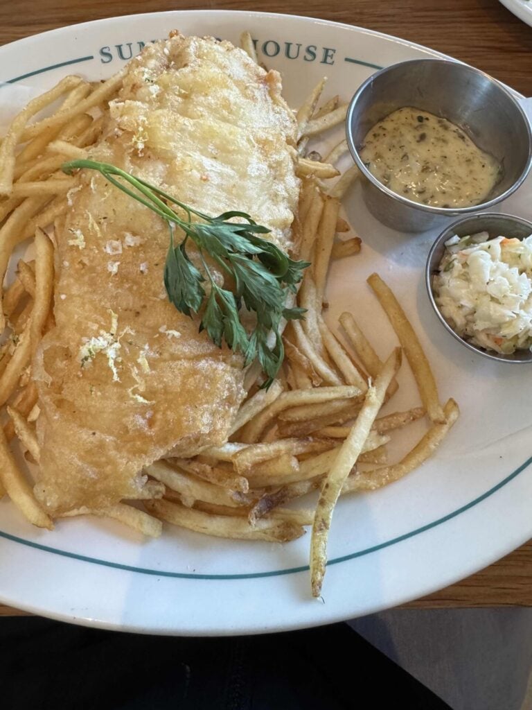 A plate of fish and chips with a sprig of parsley on top, served with a side of tartar sauce and coleslaw in small metal cups on a white plate.