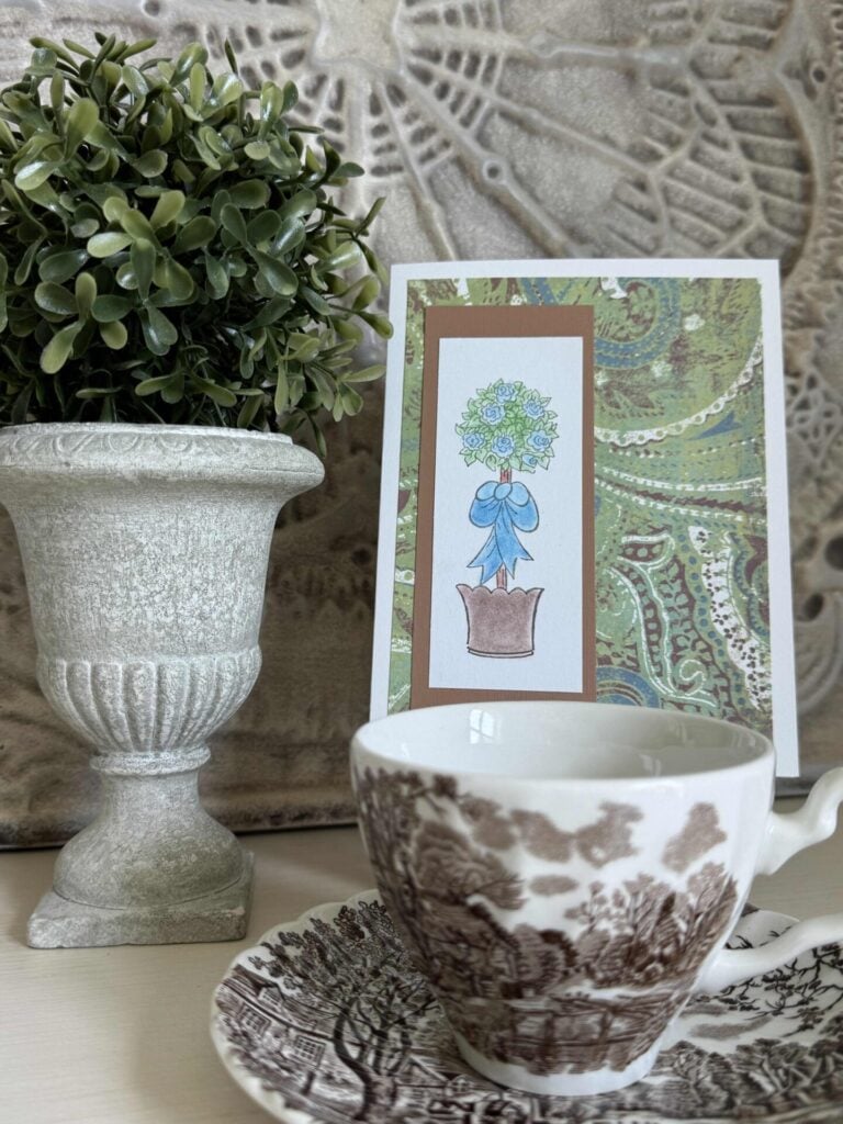 A handmade card with a topiary in green, blue, and brown next to a faux topiary and a brown transferware teacup and saucer. 