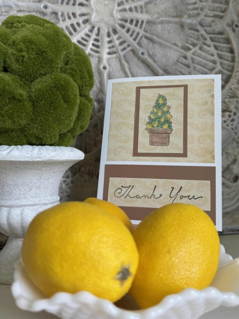 A handmade card that says "thank you" next to a faux topiary and a white bowl of lemons. 