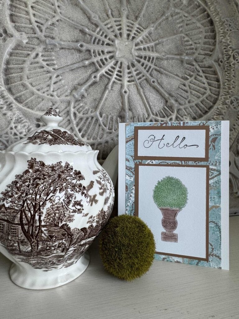 A handmade card that says "hello" by a brown transferware teapot and a moss ball. 