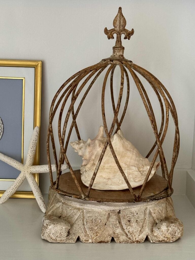 A large seashell sits under a rustic, dome-shaped metal cage with a fleur-de-lis top. Embracing coastal decor, a starfish and framed gray artwork rest nearby on the light-colored surface.