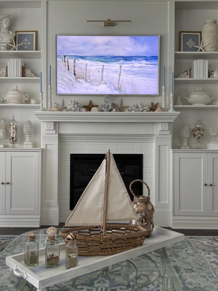 A coastal-themed living room features Coastal Decor with a white fireplace, built-in shelves displaying seashells, a TV showing a beach scene, and a coffee table adorned with a decorative sailboat and bottles filled with sand and shells.