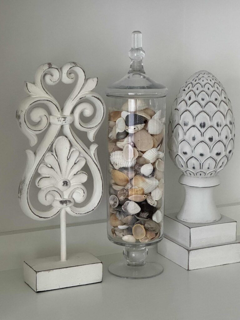 Three decorative objects on a shelf: a white ornate finial, a glass jar filled with assorted seashells, and a white artichoke-shaped ornament—all with a distressed, vintage look that brings coastal decor charm to your space.