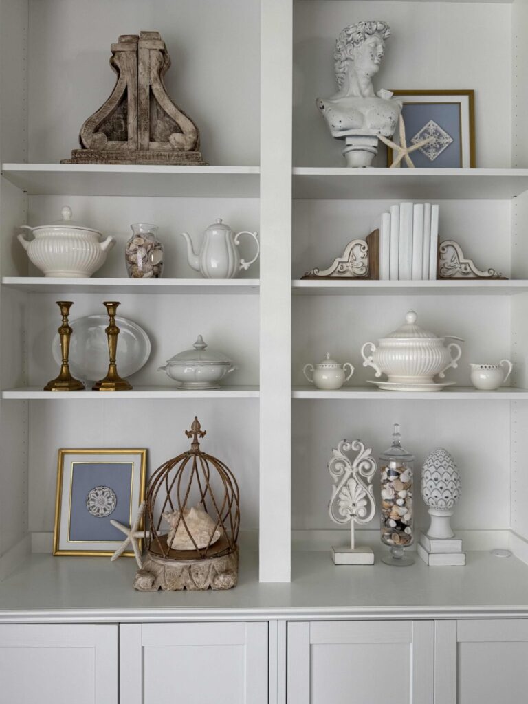 White built-in shelves display Coastal Decor: white ceramic teapots, dishes, and jars, brass candlesticks, books, framed art, shells in jars, starfish, a bust, and various vintage and coastal-themed objects.