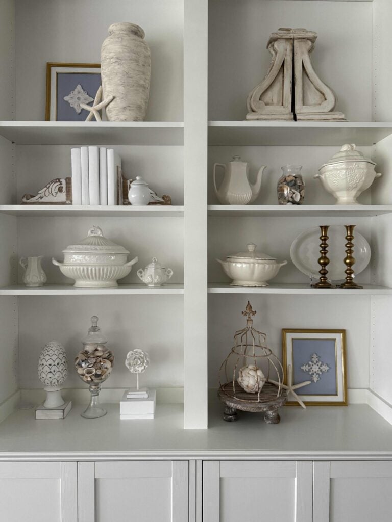 White built-in shelves display ceramic vases, teapots, tureens, glass jars with seashells, candlesticks, framed art, and decorative objects in neutral tones for elegant and organized coastal decor that exudes relaxed sophistication.