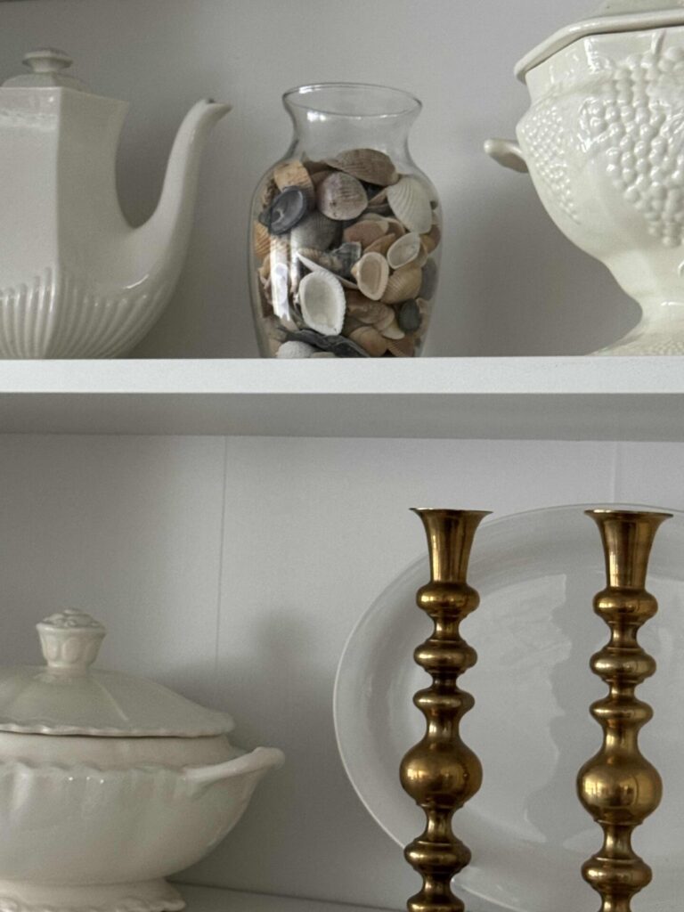 A white shelf displaying coastal decor: a white teapot, a vase filled with assorted seashells, a textured white dish, a covered white tureen, a white plate, and two gold candlesticks.
