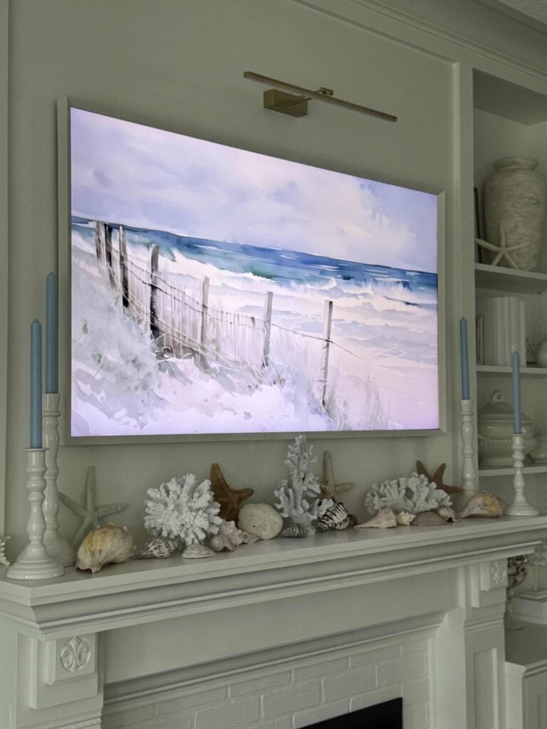 A mantel decorated with seashells, coral, and blue candles reflects coastal decor, sitting below a painting or TV display of a beach scene with waves and sand. Built-in shelves with decor are visible on the side.
