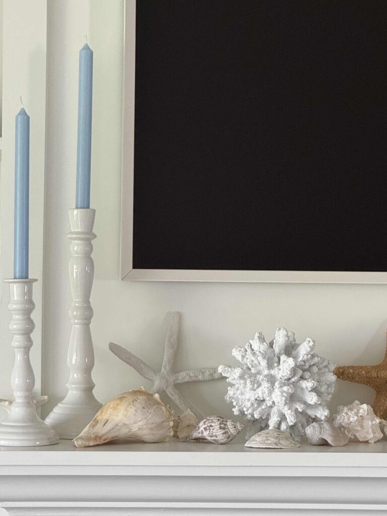 A mantel decorated with coastal decor—two white candlesticks holding light blue candles, assorted seashells, a white coral, and a starfish—all set against a white-framed black background.