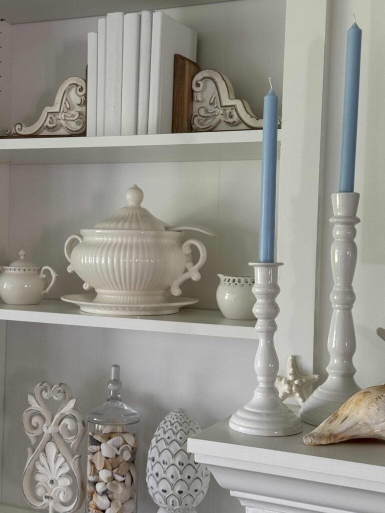 White shelves displaying decorative items: blue candles in white holders, a white ceramic tureen, teapot, and small jar, white books, ornate accents, seashells, and a large shell—perfect touches for refined Coastal Decor.