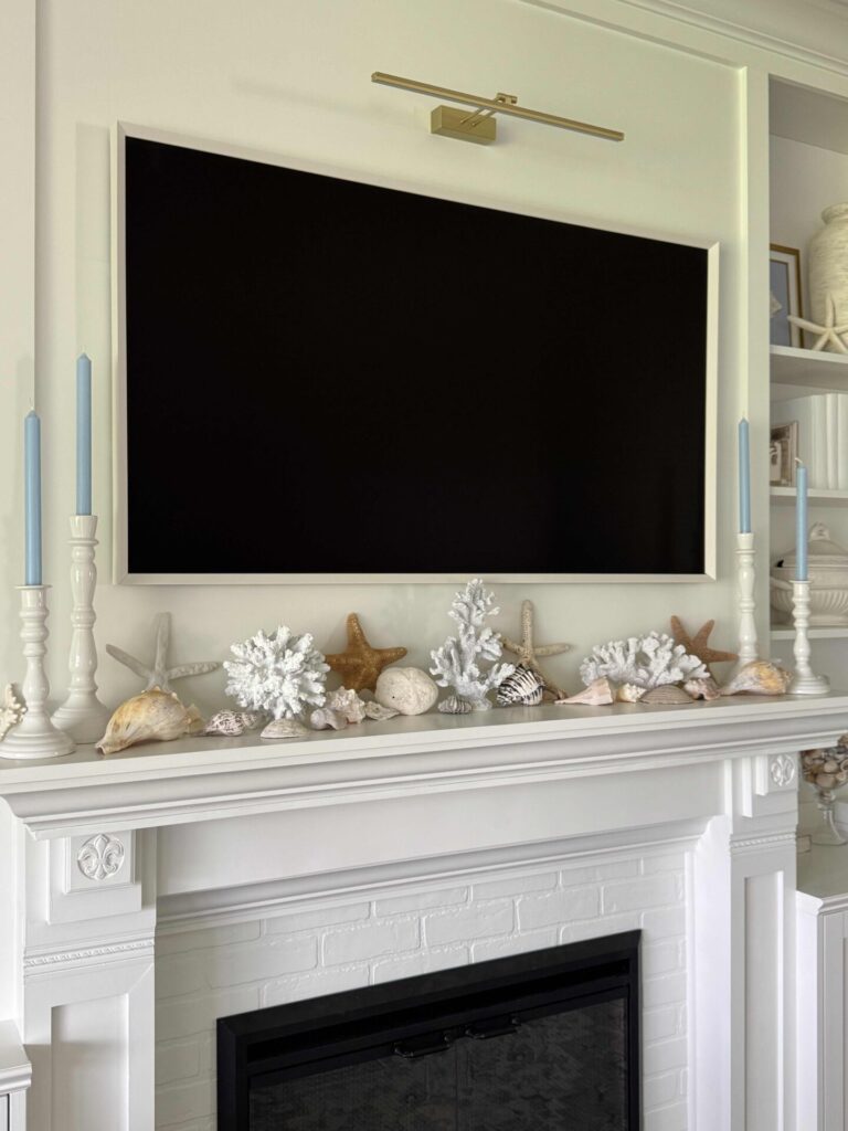 A white fireplace mantel with coastal decor like seashells, coral, and starfish. Two tall white candle holders with blue candles stand at each end. Above is a large wall-mounted TV illuminated by a gold picture light.