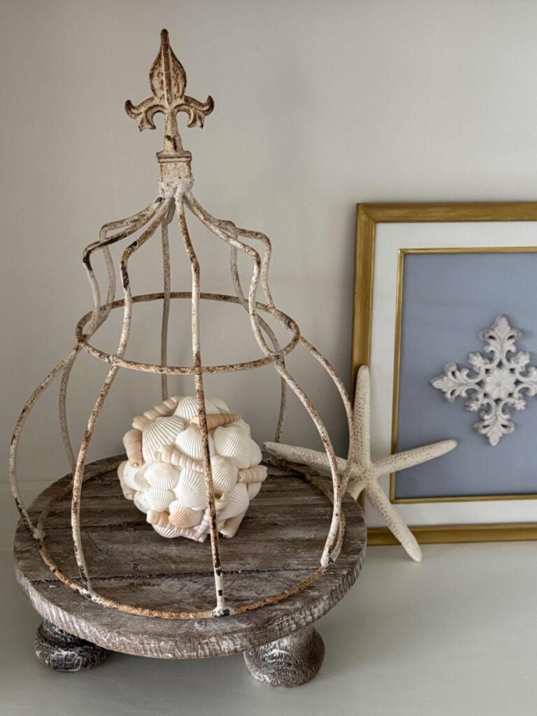 A rustic metal cloche sits on a wooden pedestal with a decorative ball made of seashells inside. A white starfish leans against a framed artwork featuring a white ornamental design in the background.