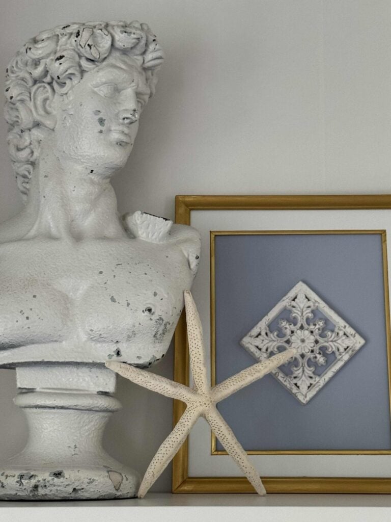 A white, weathered bust sculpture is displayed next to a large starfish and a framed decorative tile with a gold border, creating elegant coastal decor on a white shelf against a light gray wall.
