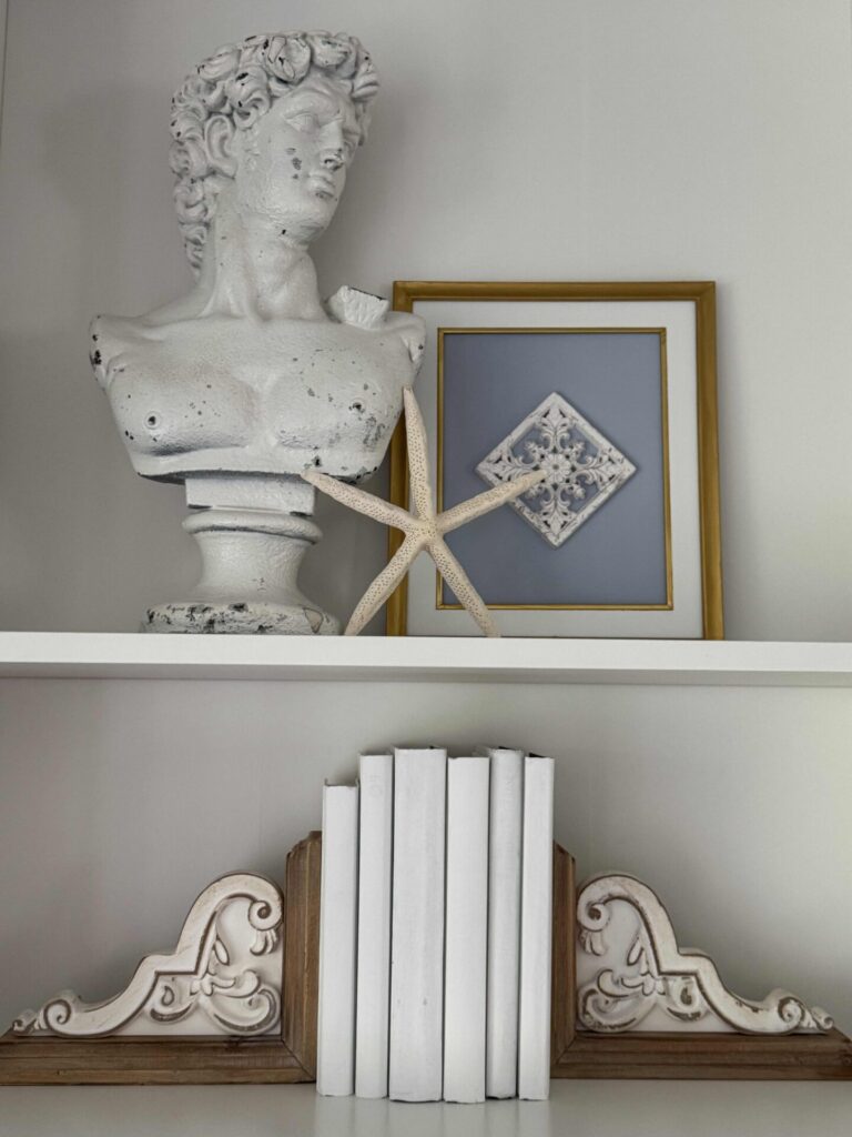A white shelf displays a classical-style bust, a starfish, and a framed art piece above another shelf with white books held by ornate wooden bookends. The minimalist and artistic decor evokes a sense of refined coastal decor.