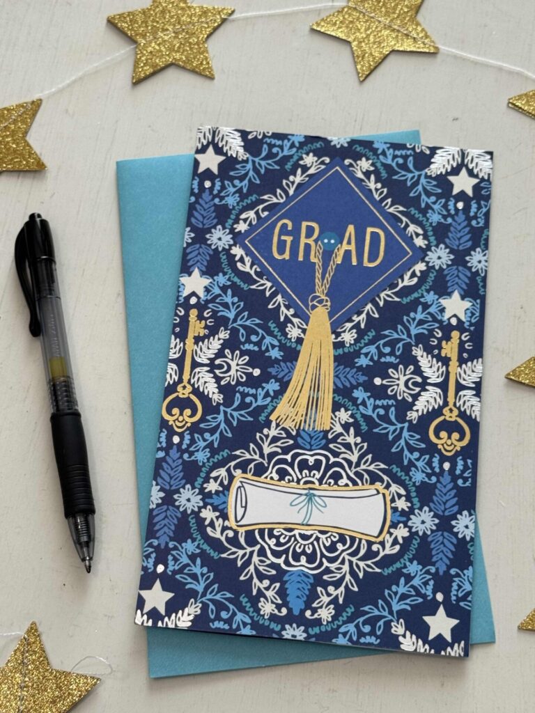 A graduation card with a blue and gold design, featuring a graduation cap, tassel, and diploma, sits on top of a blue envelope. Organize greeting cards like this with a black pen and gold glitter star decorations nearby.