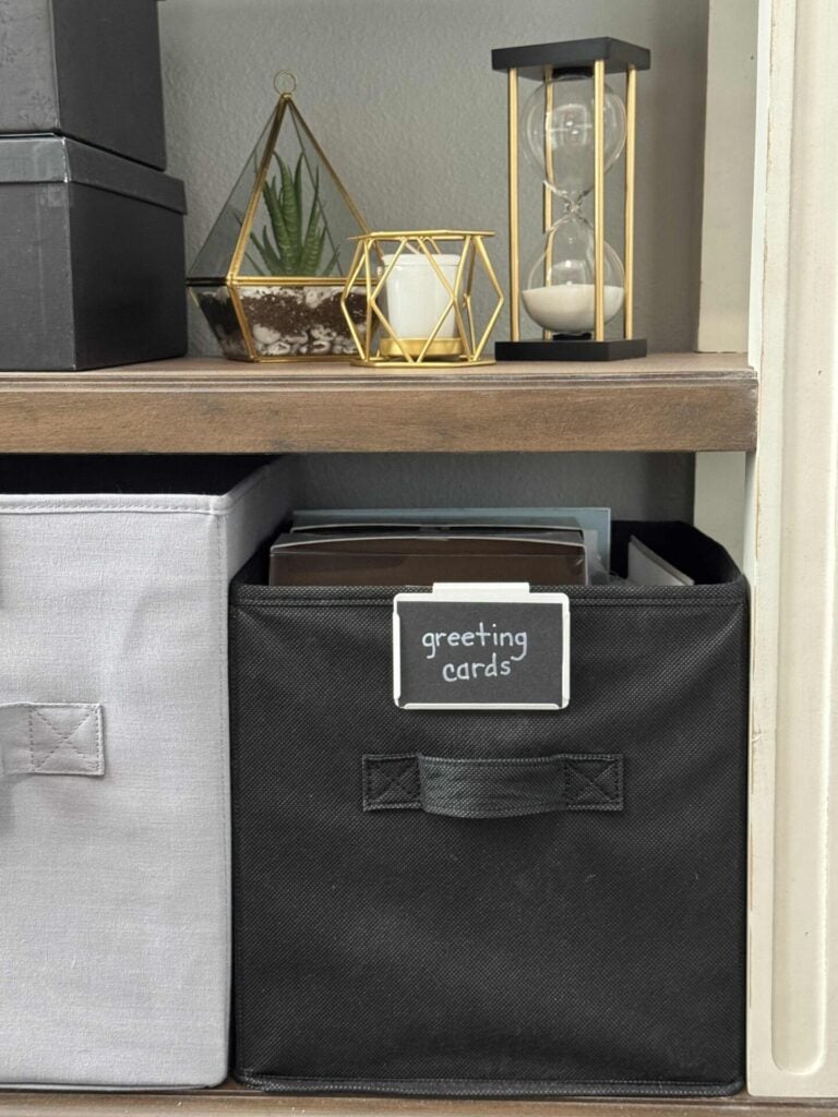 A shelf with decorative items, including an hourglass, gold geometric candle holder, and a small plant on the upper shelf, and a labeled black storage bin to help organize greeting cards on the lower shelf.