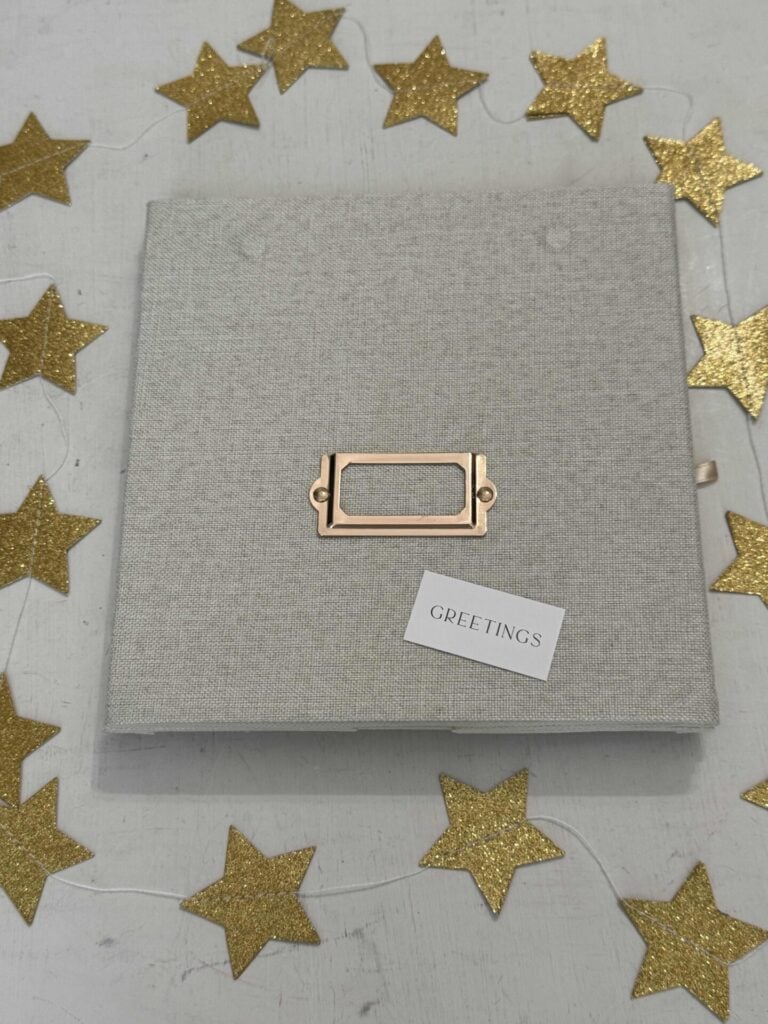 A square gray greeting card with a gold nameplate sits on a white surface, surrounded by scattered gold glitter star decorations. Organize greeting cards easily with the included small white "GREETINGS" card resting on top.