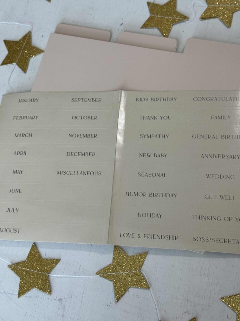 A beige folder organizer with labeled tabs and a sheet of labels for months, occasions, and themes—perfect to organize greeting cards for birthdays, sympathy, thank you, and love. Gold glittery stars are scattered on the white surface.