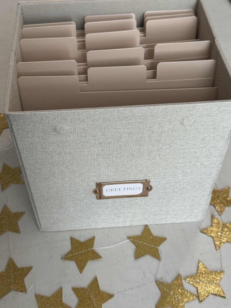 A fabric-covered box labeled GREETINGS helps organize greeting cards with neatly arranged beige dividers. Gold star-shaped decorations are scattered on the white surface around the box.