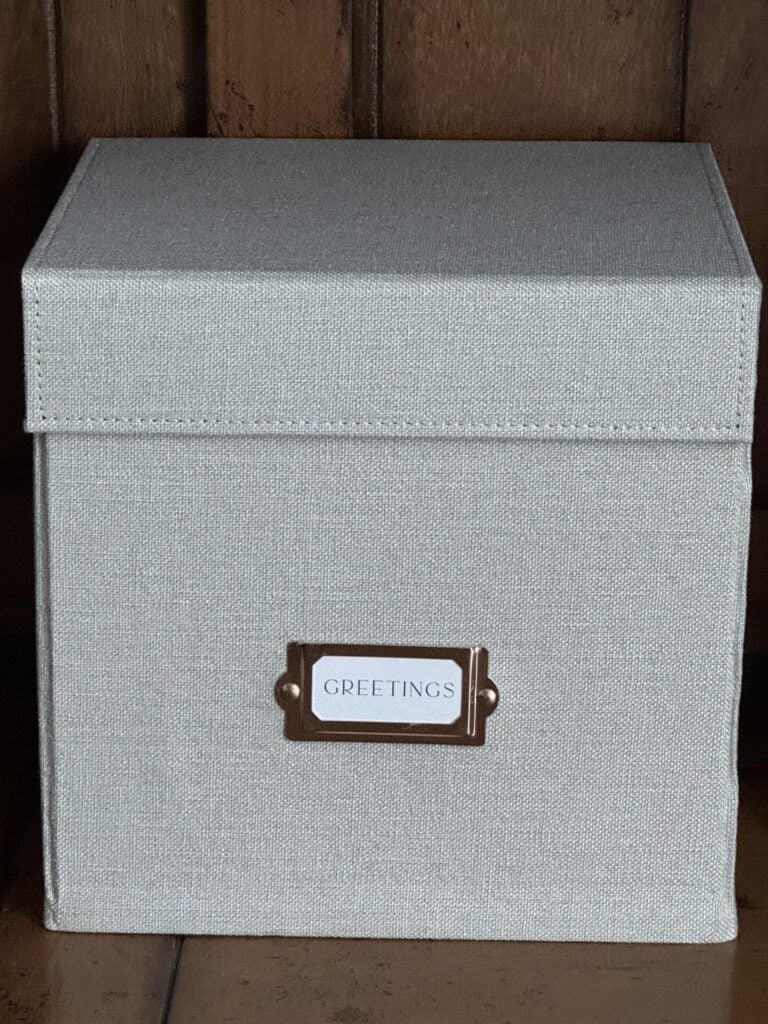 A light gray fabric-covered box with a lid sits on a wooden surface. A metal label holder on the front displays a card that reads GREETINGS, making it easy to organize greeting cards stylishly and keep them within reach.