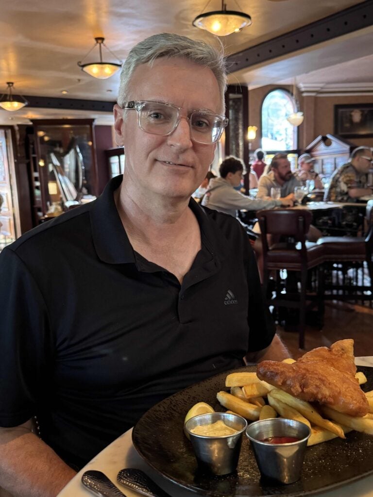 A man with gray hair and glasses, wearing a black polo shirt, sits at a restaurant table with a plate of fish and chips, sauces, and fries in front of him. Other diners are visible in the warmly lit background.