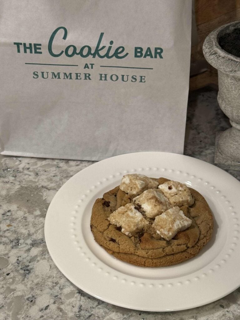 A chocolate chip cookie topped with powdered sugar-covered squares sits on a white plate. In the background, there is a bag labeled The Cookie Bar at Summer House.