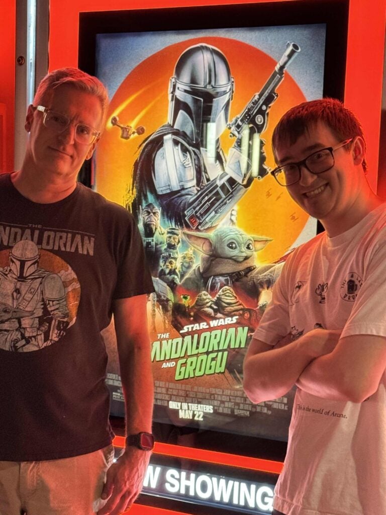 Two people stand smiling in front of a brightly lit movie poster for The Mandalorian and Grogu. The person on the left wears a Mandalorian-themed shirt; both appear excited for the film.