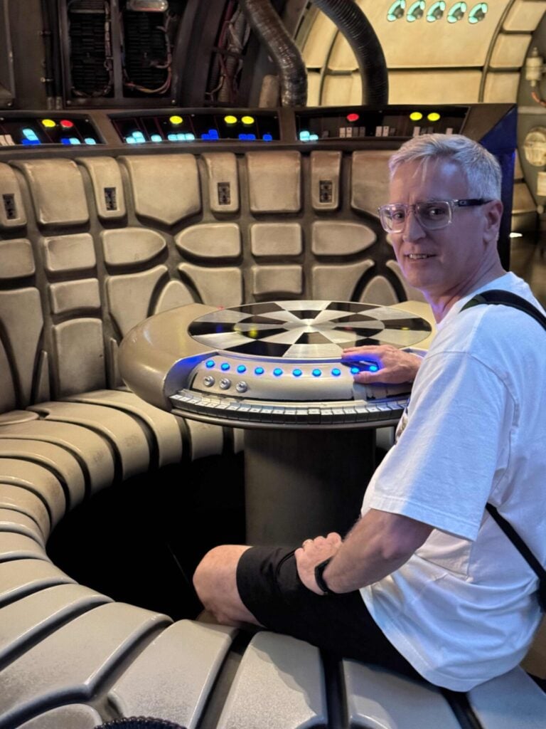 A man in a white shirt and glasses sits at a futuristic, circular control panel with colored buttons in a sci-fi themed room, smiling at the camera.