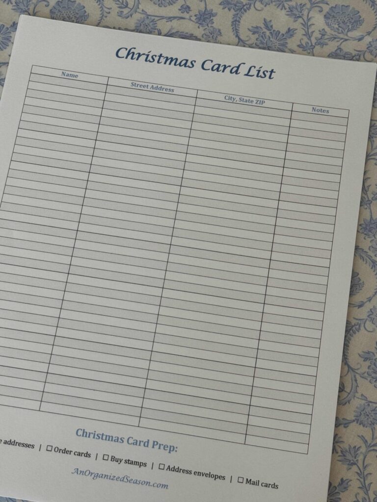 A blank Christmas Card List worksheet to help organize greeting cards, with columns for name, address, and notes. At the bottom are checkboxes for card prep tasks. The sheet rests on a blue patterned surface.