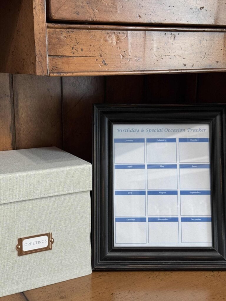 A framed Birthday & Special Occasion Tracker calendar sits on a wooden desk next to a light gray box labeled GREETINGS, helping you organize greeting cards. The calendar has blank spaces for each month.