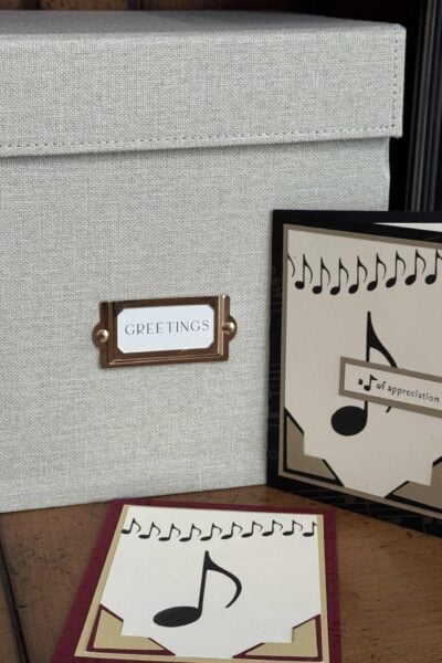 A fabric-covered greeting card storage box labeled GREETINGS helps you organize greeting cards. It sits on a wooden surface beside two handmade cards decorated with musical notes and themed designs.