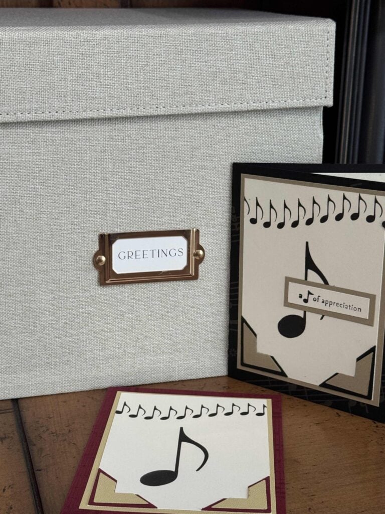 A fabric-covered greeting card storage box labeled GREETINGS helps you organize greeting cards. It sits on a wooden surface beside two handmade cards decorated with musical notes and themed designs.