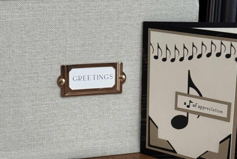 A fabric-covered greeting card storage box labeled GREETINGS helps you organize greeting cards. It sits on a wooden surface beside two handmade cards decorated with musical notes and themed designs.