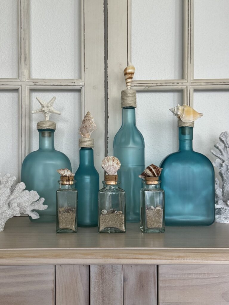A collection of blue and clear glass bottles with corks, some topped with seashells, is displayed on a wooden surface in front of a window frame. White coral pieces decorate each side, creating charming coastal decor.