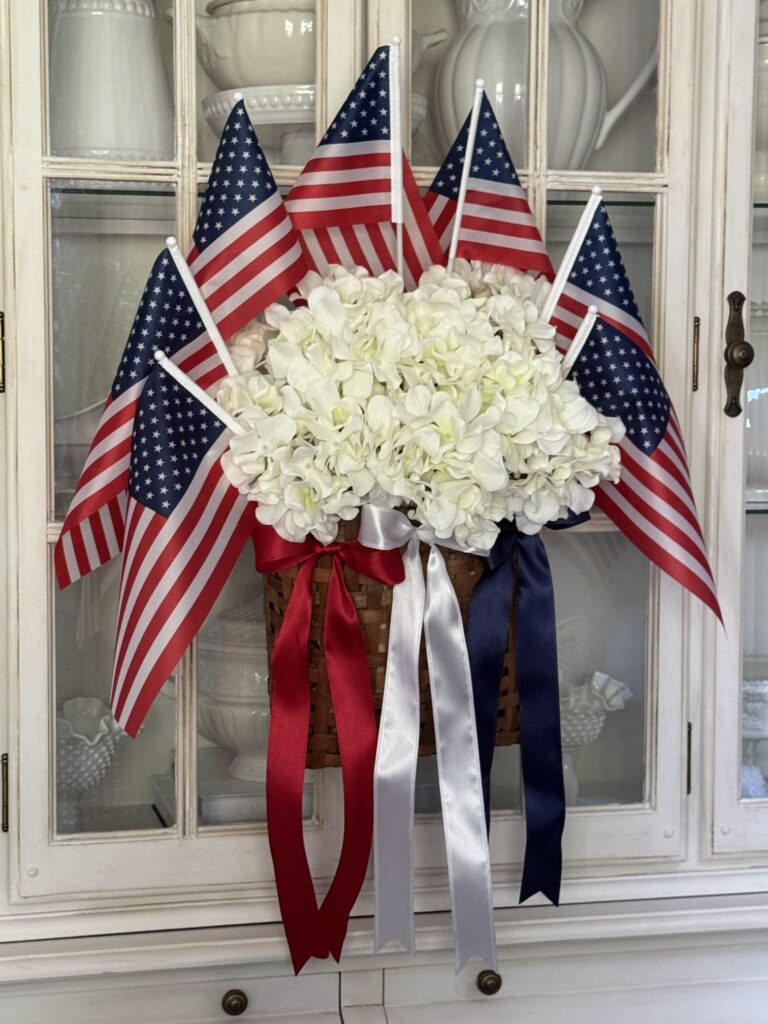 A basket of white flowers decorated with several small American flags and three red, white, and blue ribbons is displayed in front of a white cabinet—perfect for inspiring Patriotic Entertaining Ideas.