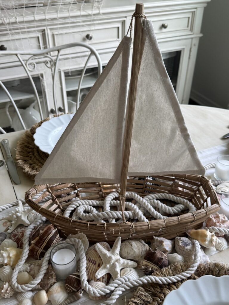 A woven decorative sailboat with a fabric sail sits on a table adorned with seashells, starfish, rope, and candles, creating a coastal-themed centerpiece—perfect for patriotic entertaining ideas. White plates and wicker placemats are set nearby.